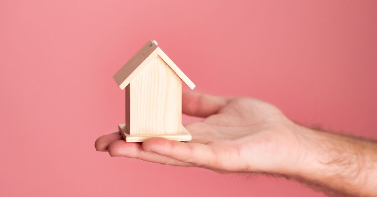 Hand holding a small wooden house model against a soft pink background, symbolizing real estate and property investment.